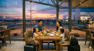 Gia đình trẻ vui vẻ ăn uống tại nhà hàng gần cửa sổ nhà ga T3 Tân Sơn Nhất lúc hoàng hôn.